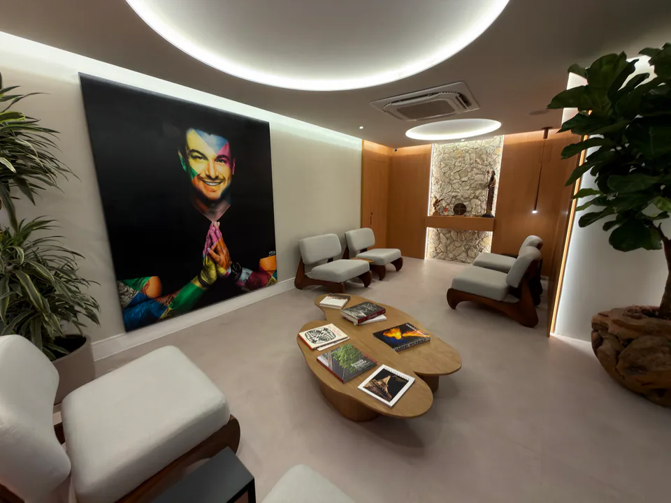 Sala de espera vista entre poltronas claras, mesas de madeira, plantas e retrato de Rafa Puglisi
